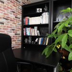 レンガの壁に囲まれたオフィスコーナーを示しており、黒いオフィスチェアとデスクの上に本棚があり、その棚にはカメラと写真撮影に関する書籍が並んでいます。室内植物が自然な雰囲気を加えています